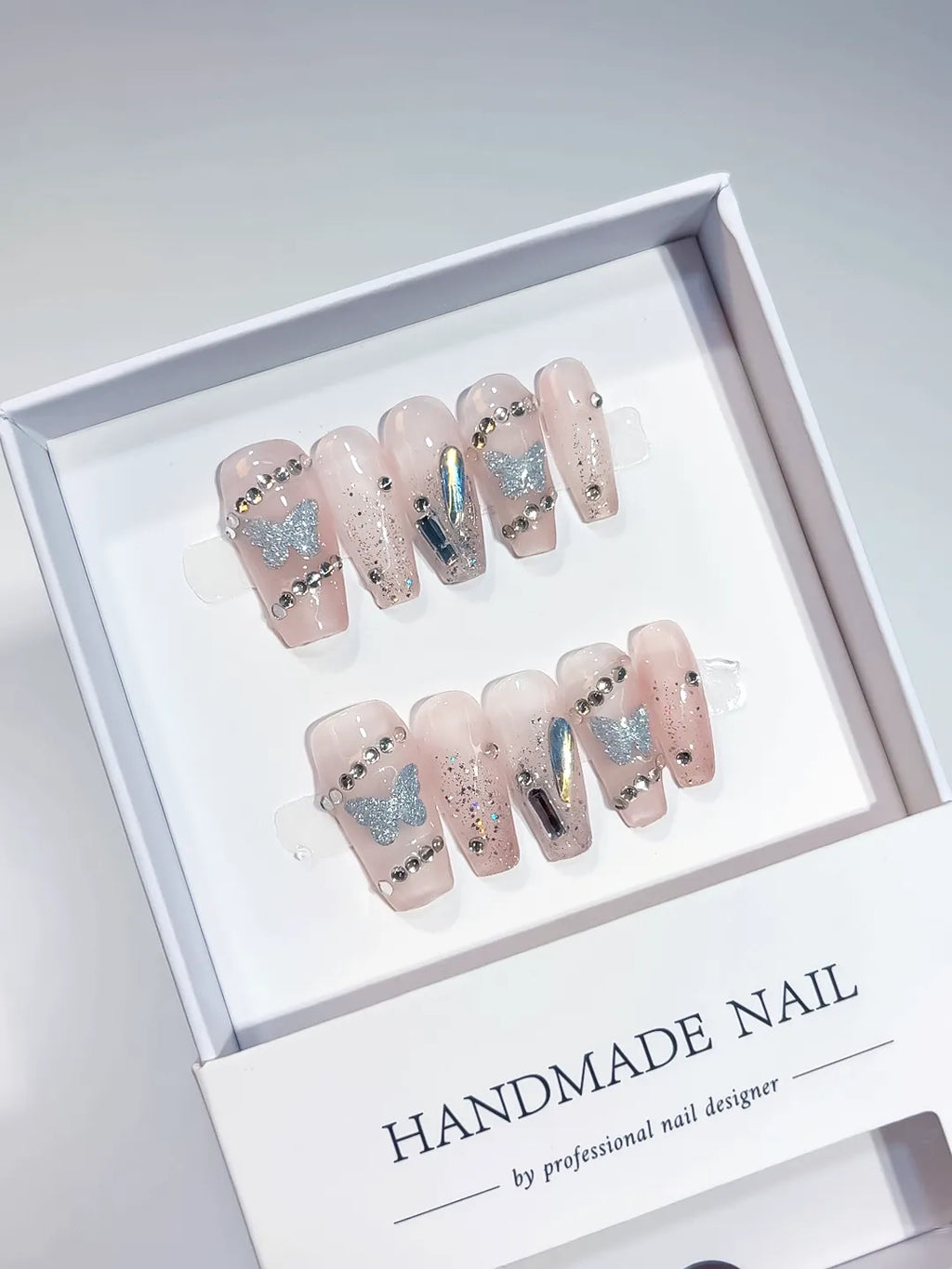 Elegant Nude Coffin Medium Handmade Press On Nails, featuring shimmering silver glitter butterflies, dazzling rhinestones, and multicolor holographic shards. This design creates a luxurious, sparkling look, perfect for a sophisticated yet whimsical aesthetic. Ideal for retail display.