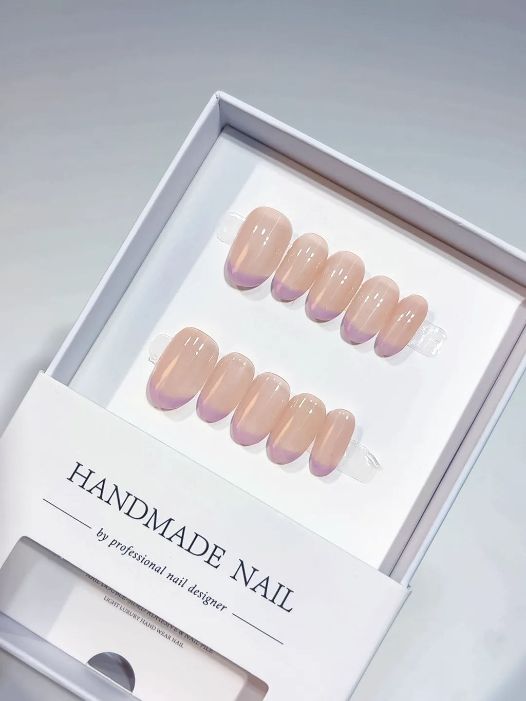 Short oval press-on nails feature a sheer nude base with soft lavender purple French tips, showcasing a glossy finish. The elegant set is displayed neatly in a clean white product box.
